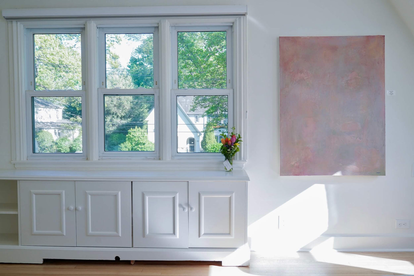 White window seat with a view of greenery, a vase with flowers, and a painting on a white wall.