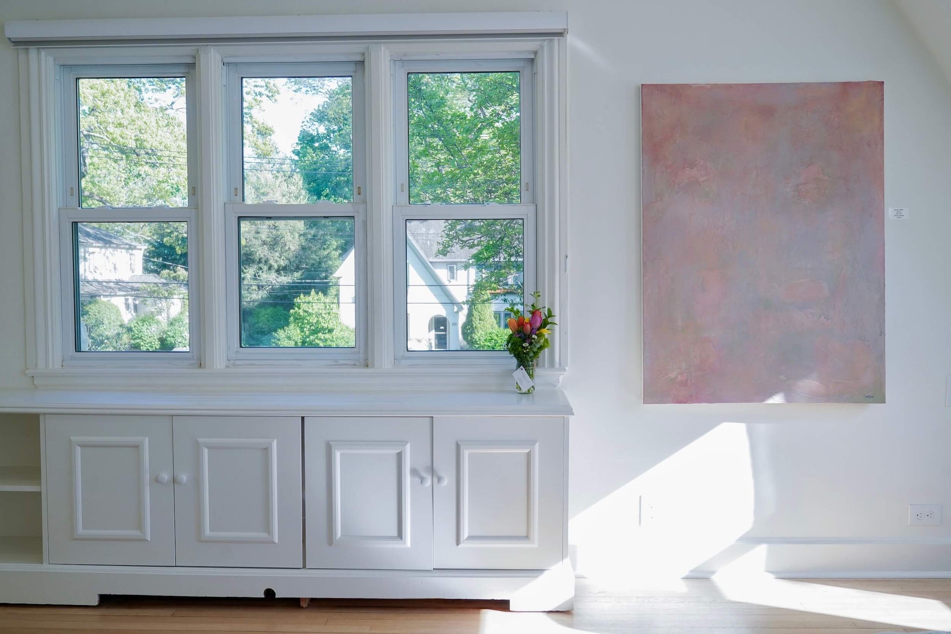 White window seat with a view of greenery, a vase with flowers, and a painting on a white wall.